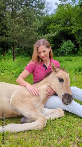 Young woman cuddling with cute small horse foal lying on grass ground, stroking fur over its head, vertical video