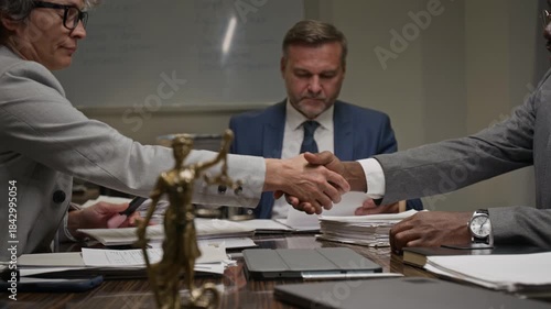 Medium shot of diverse male and female lawyers reviewing case documents at team meeting at legal firm, agreeing on contract clause and shaking hands, Lady Justice figurine on table in foreground
