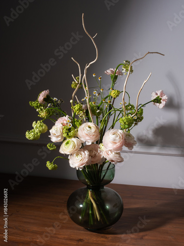 An elegant bouquet features pale pink ranunculus, carnations, chamomile, green spirea and bare branches in an olive green bubble vase on a dark wooden table. Gray wall background.