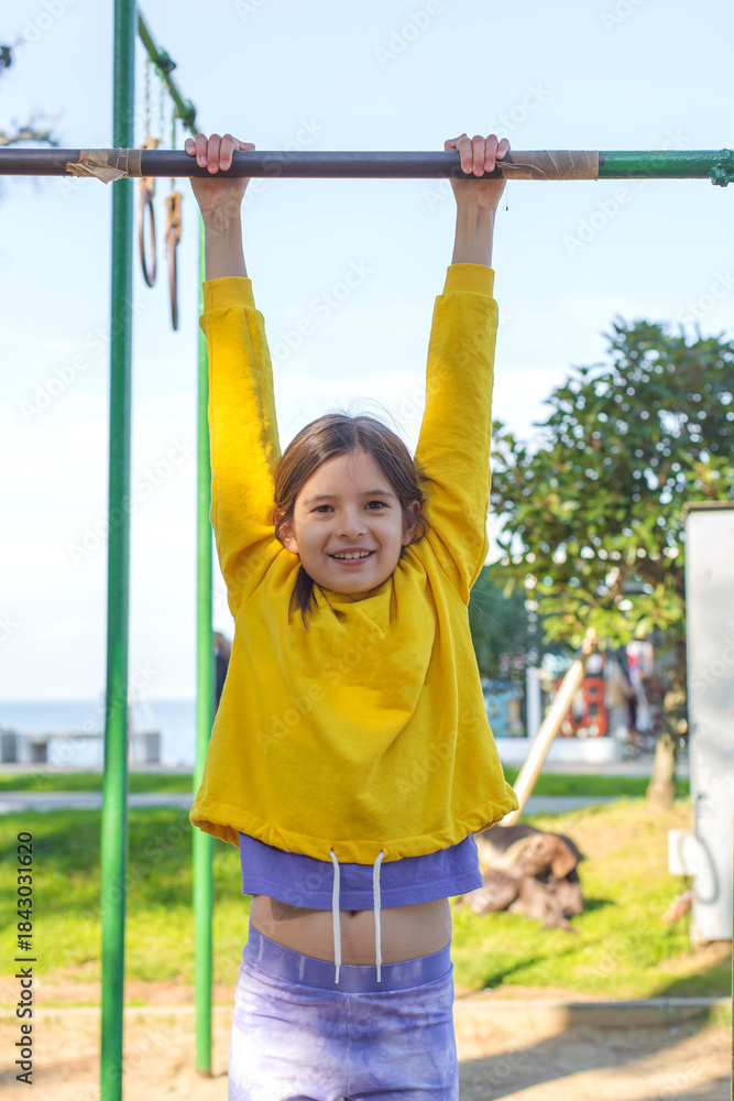 Fototapeta premium Smiling girl hanging on outdoor bar during active play, enjoying sunny weather and practicing strength and coordination in a park near the seaside, creating a bright and energetic childhood moment