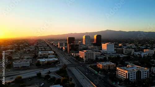 Aerial view of a modern city skyline with tall skyscrapers and mountains in the background during a vibrant golden hour sunset.