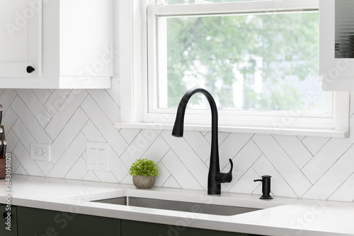 Sink detail shot in a luxury kitchen with white herringbone backsplash tiles. white marble countertop, white and green cabinets, and a black faucet.