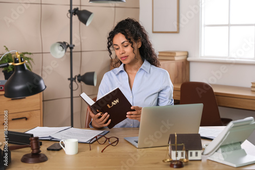 Beautiful African-American lawyer with law book at table in office