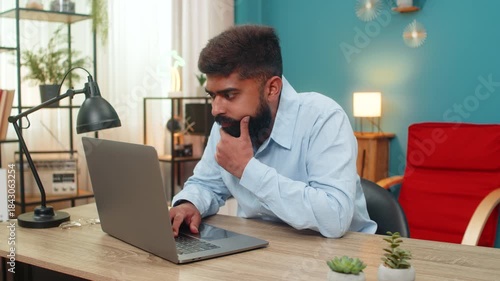 Indian man at home types on laptop stops rubs chin thinking over vital idea creative future plan. Arabian guy at table gazes screen focused eyes show deep concentration solving problem quickly today.