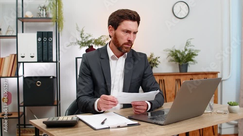 Middle-aged businessman at home office enters numbers on laptop checks amounts with calculator and receipts. Freelancer guy at table reviews bills and signs documents with calm focus and steady hand.
