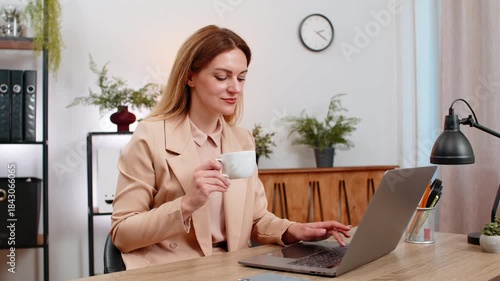 Middle-aged businesswoman at home office reads task list on laptop drinks coffee takes sip smiles stays focused. Freelancer girl at table enjoys cup gets energy plans steps types reply and saves file.