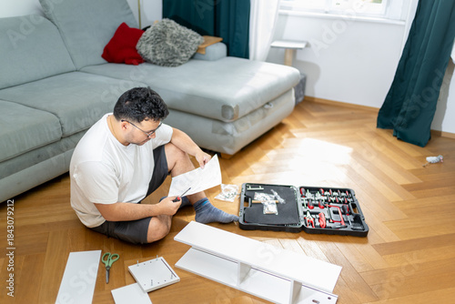 Young man assembling flat pack furniture at home reading instruction manual with tools