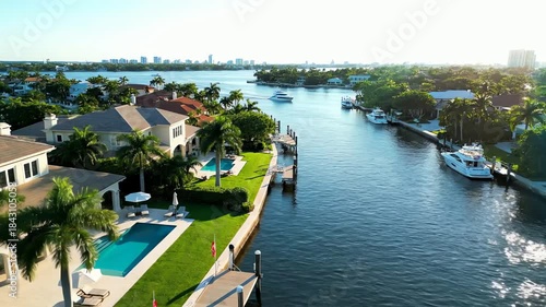Stunning aerial view of luxurious waterfront mansions with private yachts along a serene canal with a city skyline in the distance.