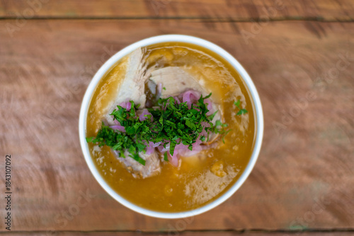 A plate of encebollado on a wooden table in the foreground, with cilantro, fish, and soup visible