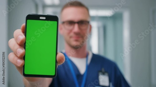 Male doctor in blue scrubs and glasses holding up a smartphone with a green screen display in a blurred hospital hallway.