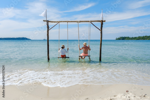 A diverse couple relaxing on swings by the serene beach of Koh Kood Island, Thailand. The calm waves and bright blue sky create a peaceful tropical atmosphere, perfect for unwinding.