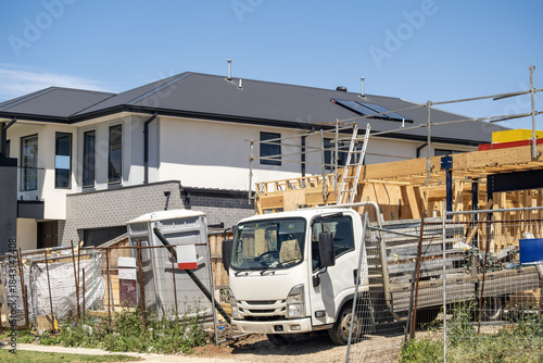 A residential construction site in a newly developed suburban estate in Australia, featuring a partially built house framed with timber, scaffolding, and safety fencing. A work truck is parked on site