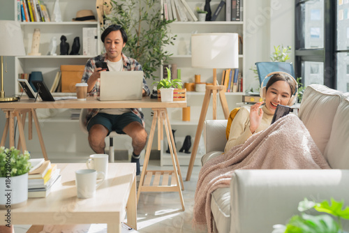 husband works remotely at home on his laptop while his wife lies cheerfully on the sofa, relaxing peacefully and enjoying a calm lifestyle moment together, showing balance, comfort, and happiness