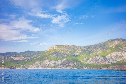 Wallpaper Mural Wide-angle seascape featuring layered green and rocky mountains along a tranquil coastline, viewed across a calm deep blue sea under a bright sky with soft clouds Torontodigital.ca