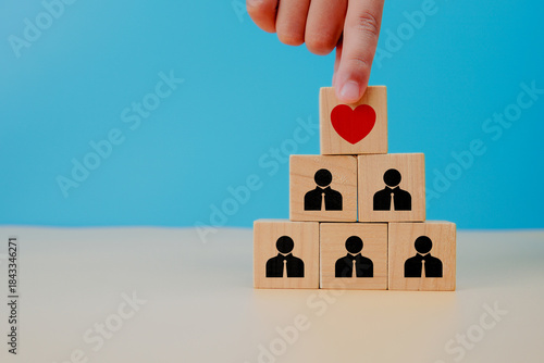 Leadership and employee wellbeing concept. Hand placing heart icon above people symbols on wooden cubes, representing care, empathy, support, motivation, and people centered management in workplace.