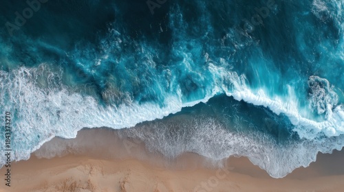 Waves crash against a sandy shore with foam forming at the water's edge. The ocean displays hues of blue and green under bright sunlight. The scene captures the movement of water and shoreline.