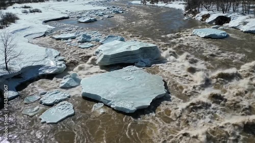 Aerial view of massive ice floes breaking up and navigating turbulent brown river water during spring thaw season