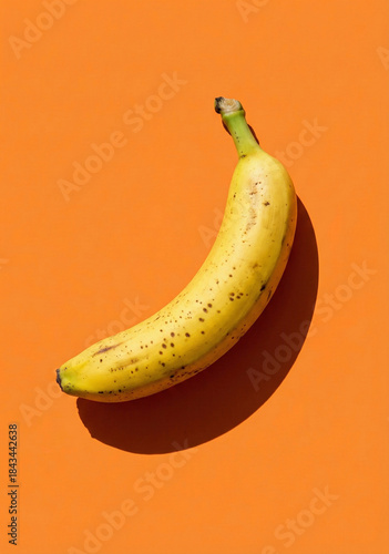 Single ripe yellow banana with brown spots on a vibrant orange background.