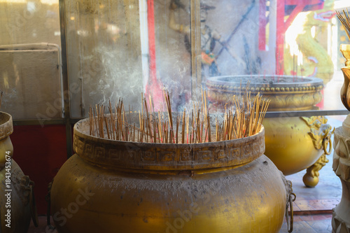 Burning incense sticks with aromatic smoke rising from large traditional golden urn at chinese temple, representing faith, prayer, and religious worship in asian culture.
