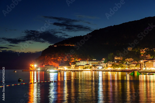 Fototapeta Naklejka Na Ścianę i Meble -  sunset in small resort town Petrovac, Montenegro, beautiful sea view, city embankment with street lights, beach and mountains against sunset sky