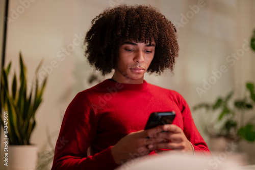 A young person with curly hair wearing a red sweater is focused on their smartphone, holding it with both hands.