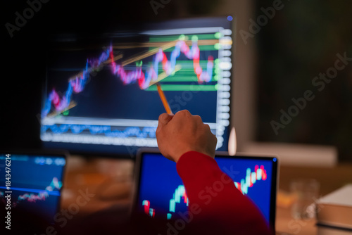 A person in a red shirt uses a pencil to point at a stock market graph on a computer screen, with other screens showing similar data in the foreground.