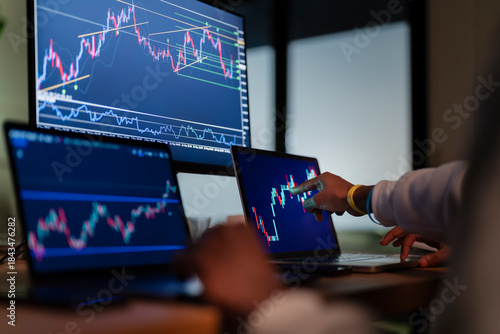 A person's hand points to a stock market chart on a laptop screen, with other monitors displaying financial data in the background.