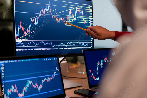 A person points at a stock market chart on a computer screen, analyzing financial data with multiple monitors displaying trading graphs.
