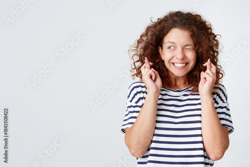 Closeup of happy lovely young woman with curly hair wears striped t shirt keeps fingers crossed and looks to the side isolated over white background Wishing for good luck