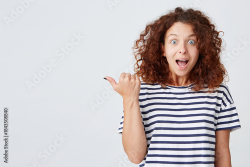 Closeup of amazed cute curly young woman with opened mouth wears striped t shirt looks surprised and points to the side with finger isolated over white background