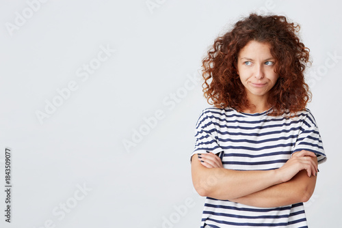 Portrait of unhappy sad curly young woman wears striped t shirt looks to the side and stands with arms crossed isolated over white background