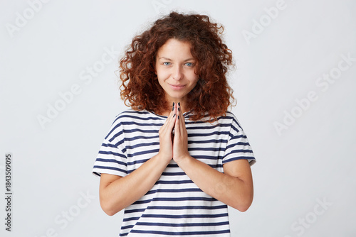 Closeup of happy peaceful young woman with curly hair and wears striped t shirt looks calm and stands with hands folded in praying position isolated over white background
