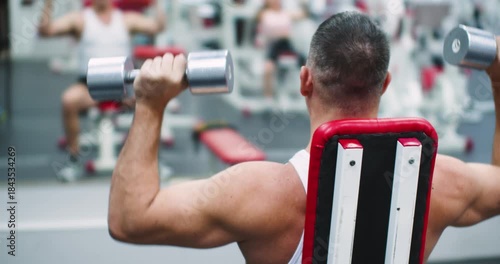 Seated bench press with dumbbells for the deltoid muscles. Deltoid workout with dumbbells. Back view of a middle aged brutal Caucasian man during gym workout in front of a mirror. 4k footage.