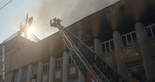 Silhouette of firefighters in a bucket on the ladder of a fire truck, extinguishing the roof of a burning building, black smoke rising, low-angle view, wide shot