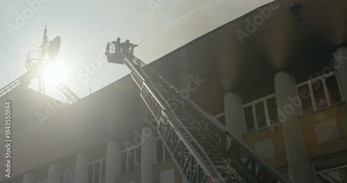 Silhouette of a fireman in a bucket on the ladder of a fire truck, extinguishing the roof of a burning building, low-angle view, wide shot