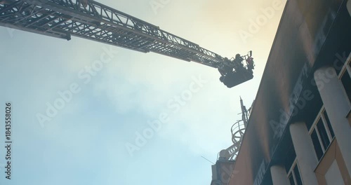 Silhouette of firefighters in a bucket on the ladder of a fire truck lifting on the roof of a burning building, low angle view.