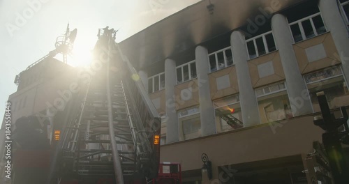 Silhouette of firefighters in a bucket on the ladder of a fire truck, extinguishing the roof of a burning building, sun through the  smoke in the background