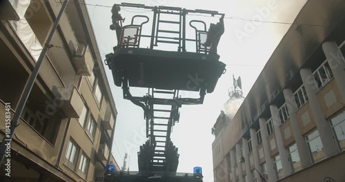 Ladder of a fire truck pulling out and lifting into the air, burning building in the background, white smoke rising.
