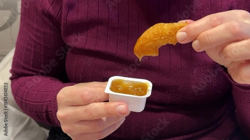 Woman Hand Holding Chicken Nugget Over Sauce, CloseUp Of Casual Eater In Maroon Shirt Dipping Golden Crispy Nugget Into HoneyMustard. Remote Worker Enjoying Quick food.