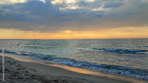 Sea waves with blue water on sand of seashore on sunset and sunrise. Yellow reflection of sunlight on surface of sea waves. Orange sky with dark rain clouds, horizon line and sea waves on sand.