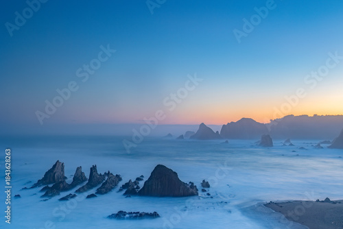 Gueirua Beach in Asturias at dawn, bathed in the first light of day. A peaceful coastal scene with dramatic rocks, soft colors, and a calm atmosphere that captures the beauty of early morning.