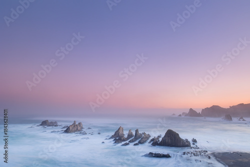 Gueirua Beach in Asturias at dawn, bathed in the first light of day. A peaceful coastal scene with dramatic rocks, soft colors, and a calm atmosphere that captures the beauty of early morning.