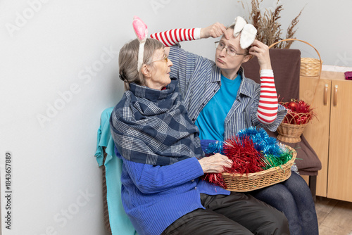 Woman social worker or daughter trying on new year bunny ears with an elderly woman, preparing for New Year and other winter holidays together, generation leisure time.