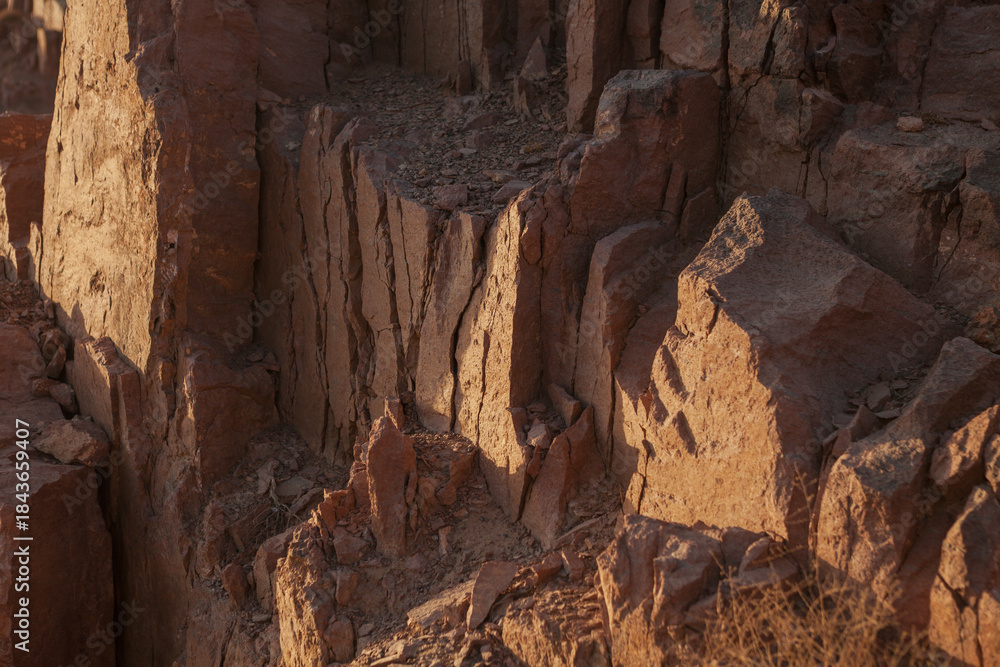Obraz premium Rocky desert cliffs near Qom, Iran, illuminated by warm sunlight. The rugged brown formations show vertical fractures and natural textures, revealing the geological layers shaped by erosion.
