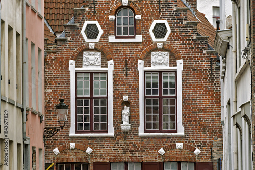 Fototapeta Naklejka Na Ścianę i Meble -  Facade of a historic brick building in Bruges featuring two large rectangular windows with white trim and red frames. Above each window are ornate white stone carvings, and a small statue stands betwe