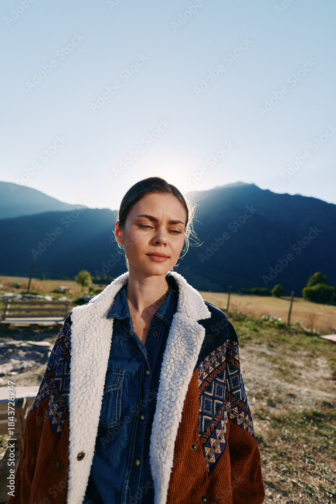 Fototapeta premium Woman portrait in mountains with sunlight behind her, eyes closed and serene expression, wearing warm patterned coat and denim shirt in outdoor nature with soft backlight.
