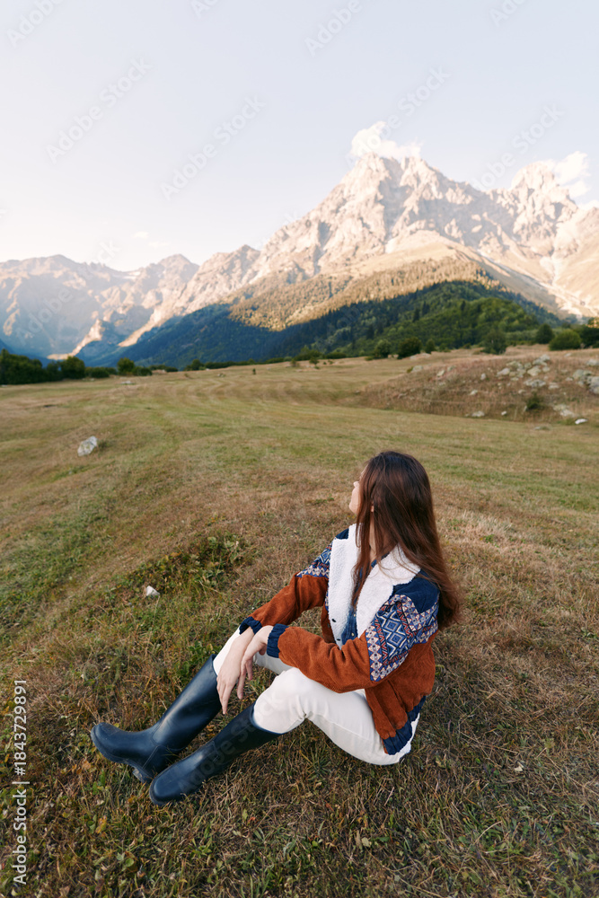 Fototapeta premium Woman sits on a meadow facing a mountain landscape during autumn travel, wearing cozy sweater and boots, enjoying serene scenery and wide grassy valley with soft sunlight.