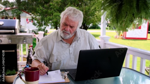 An older man sits at a table in a garden, focused on his laptop. He enjoys a cup of coffee while taking notes and working amidst green plants and a serene backdrop.