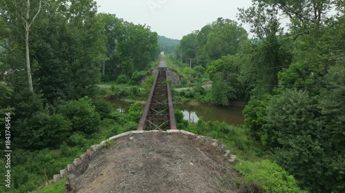 An abandoned railway bridge surrounded by vibrant trees and overgrown plants. The view highlights the lush green scenery and the structure's rustic charm, creating a serene atmosphere.