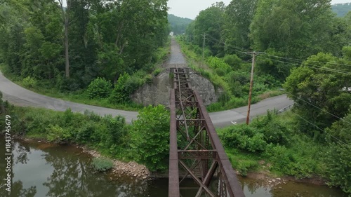 An abandoned railway bridge stretches over a calm river. Dense trees and vegetation cover the area, creating a serene and peaceful atmosphere.